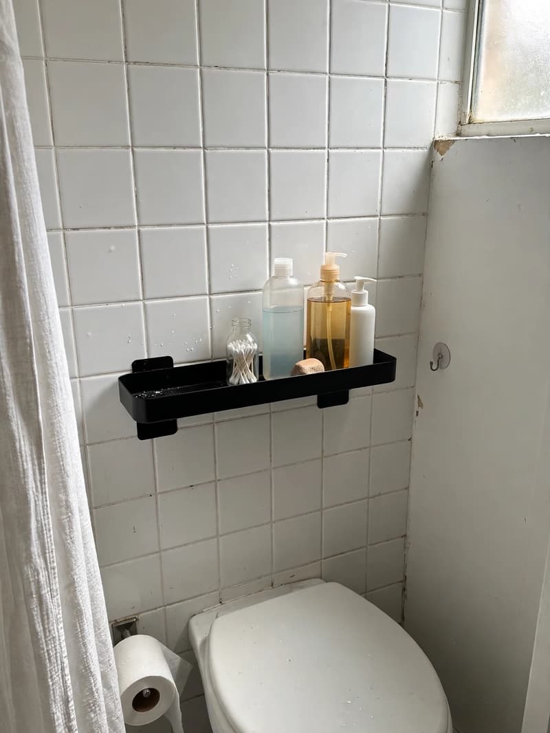A sleek black adhesive-mounted shelf on a white tiled wall above a toilet holding pump bottles and a small jar