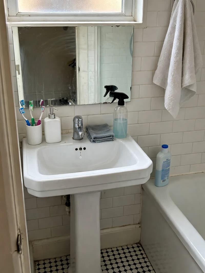 A small tiled apartment bathroom with a clean pedestal sink, spray bottle and folded cloth beside the mirror after a quick blitz