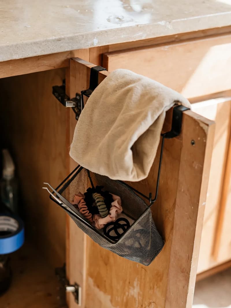 Over-the-door towel bar with a folded hand towel on a bathroom vanity door and a small hanging bin inside holding hair ties and tweezers