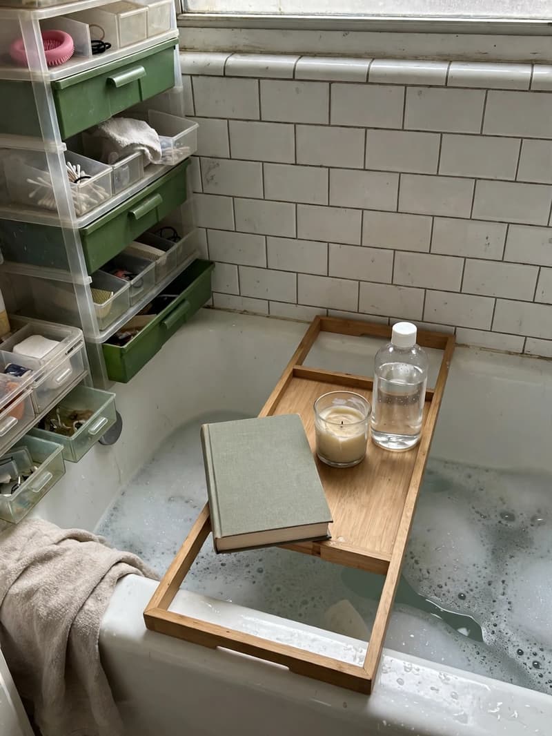 Bath tray across a bathtub holding a closed book, a small candle and a plain bottle beside the bubbles