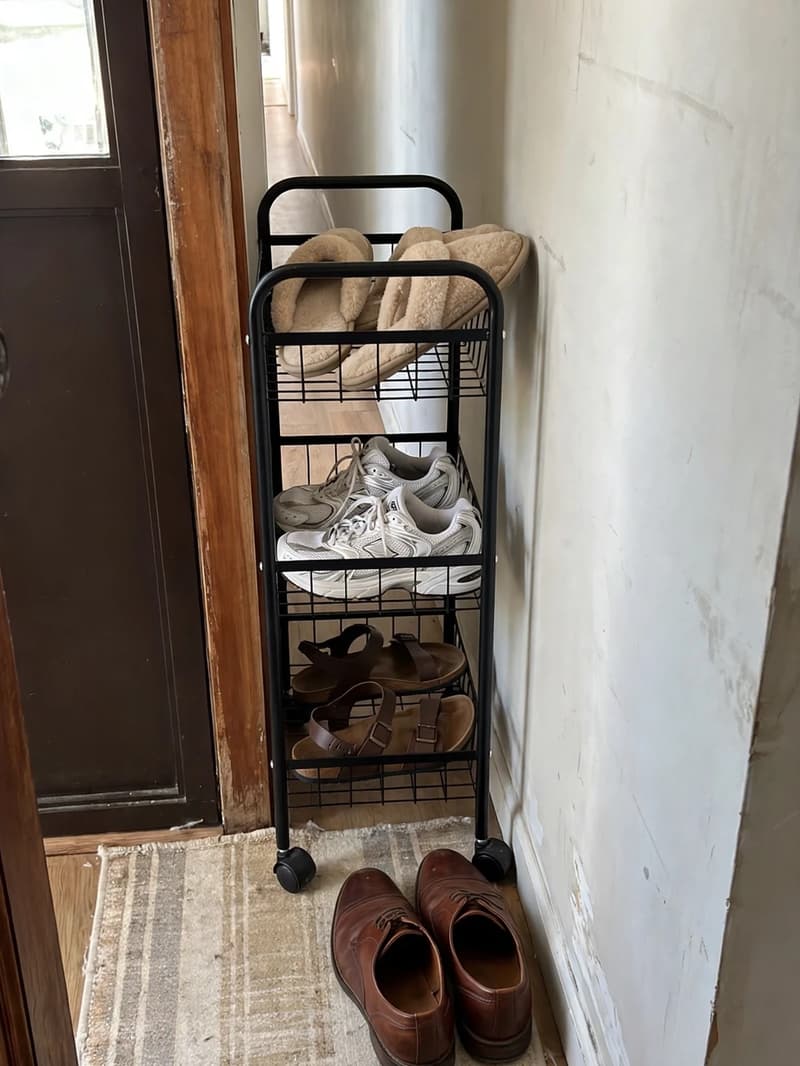 A slim black metal rolling cart with three tiers tucked into a narrow gap beside a front door frame holding slippers, sneakers and sandals