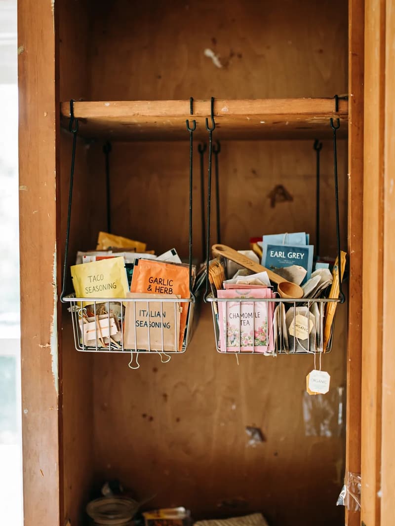 Wire under-shelf hanging baskets inside a cabinet holding seasoning packets and tea bags