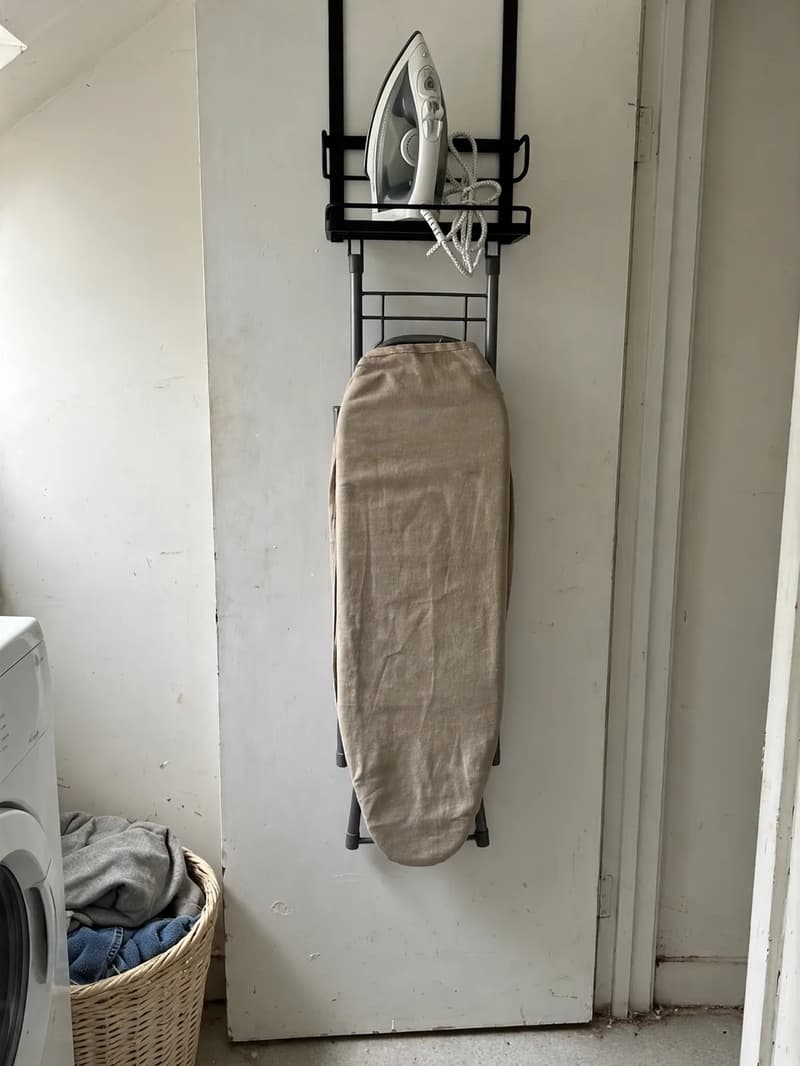 A black over-door holder on a laundry room door holding a steam iron upright above a folded ironing board hanging flat against the door