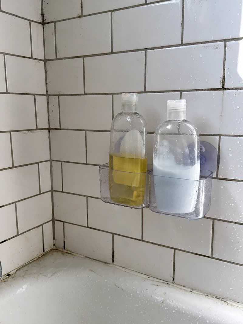 A clear adhesive shower caddy mounted on a white tiled shower wall, holding shampoo bottles above the tub edge