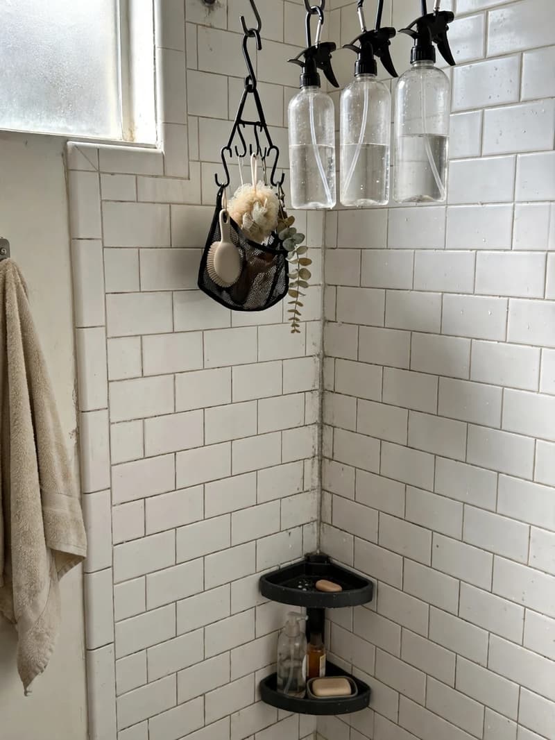 A shower corner with a tension rod holding hanging spray bottles and a small mesh caddy on hooks