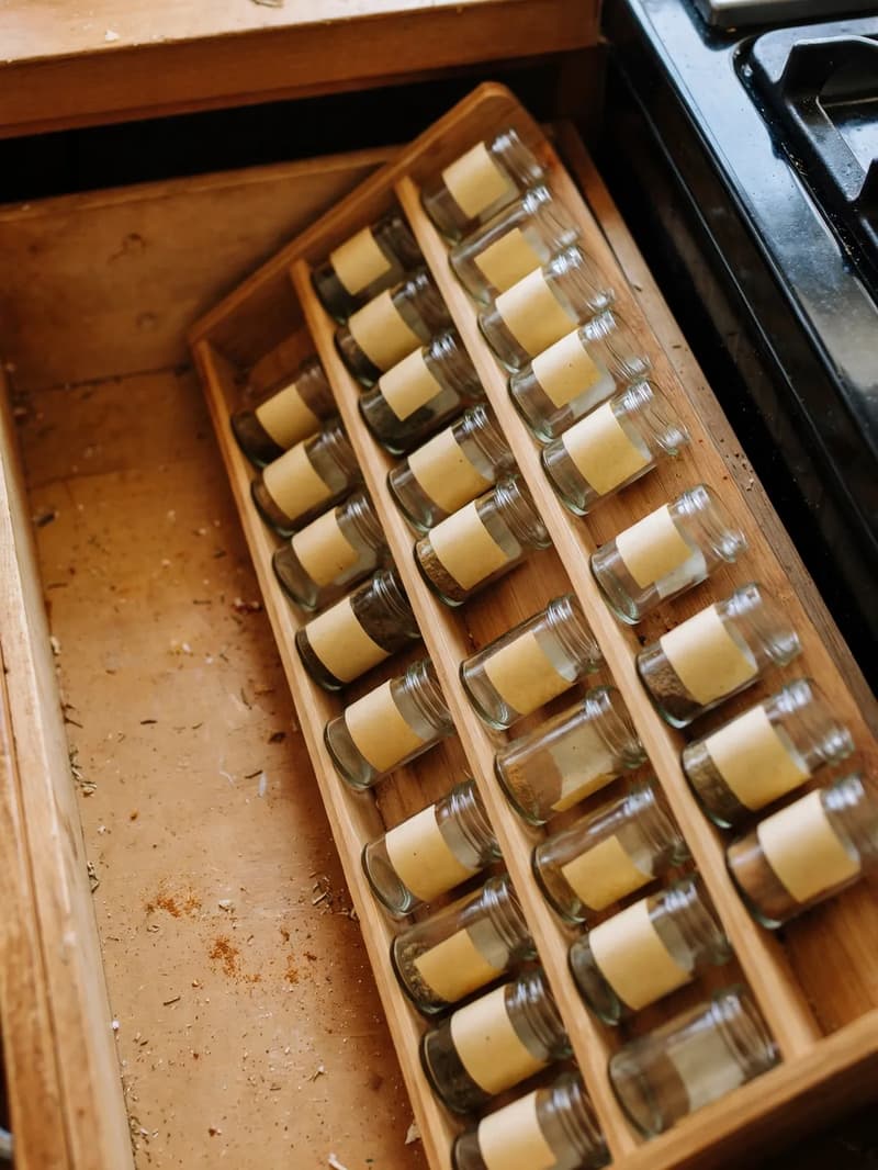 An angled tiered insert in a shallow drawer beside a stove, rows of glass spice jars lying at an angle with blank labels